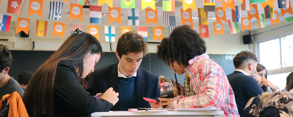Los alumnos de la UM obtienen excelentes resultados en los exámenes de chino, francés y portugués. (Foto de 2019). Tres alumnos dialogan alrededor de una mesa. Arriba se ven banderines con bandereas de distintos países.