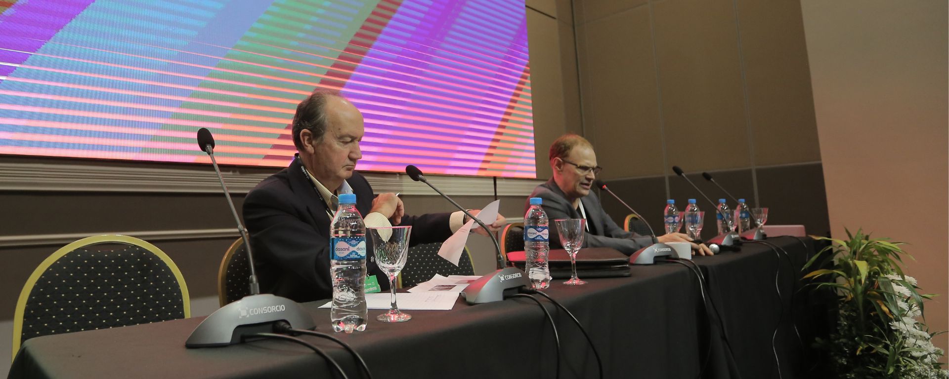 Nicolás Eycheverry en mesa de expositor junto a otro expositor, frente a micrófono (pantalla gigante detrás)