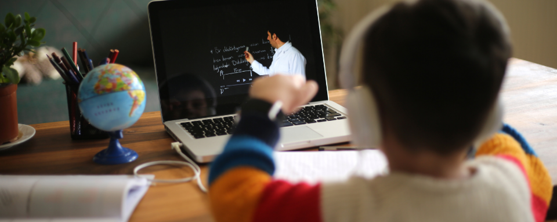 Niño de espaldas con auriculares mira hacia computadora en la que se ve un maestro dando una clase.