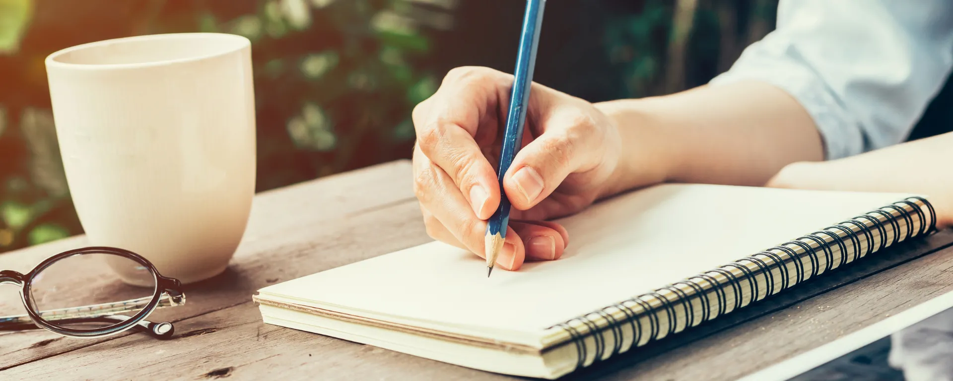 Taller de escritura: mano escribiendo con lápiz sobre cuaderno, en una mesa en exteriores, con una taza de café Taller de escritura: mano escribiendo con lápiz sobre cuaderno, en una mesa en exteriores, con una taza de café